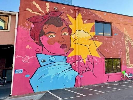 A mural of a woman in a headscarf rolling up her sleeve in front of a yellow sun, painted across a pink and coral building