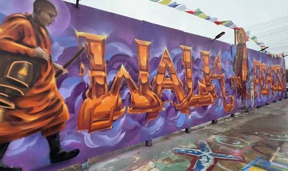 A long hand painted wall reading WALLS OF PEACE in orange lettering, with robed monk figures at each end and prayer flags strung above